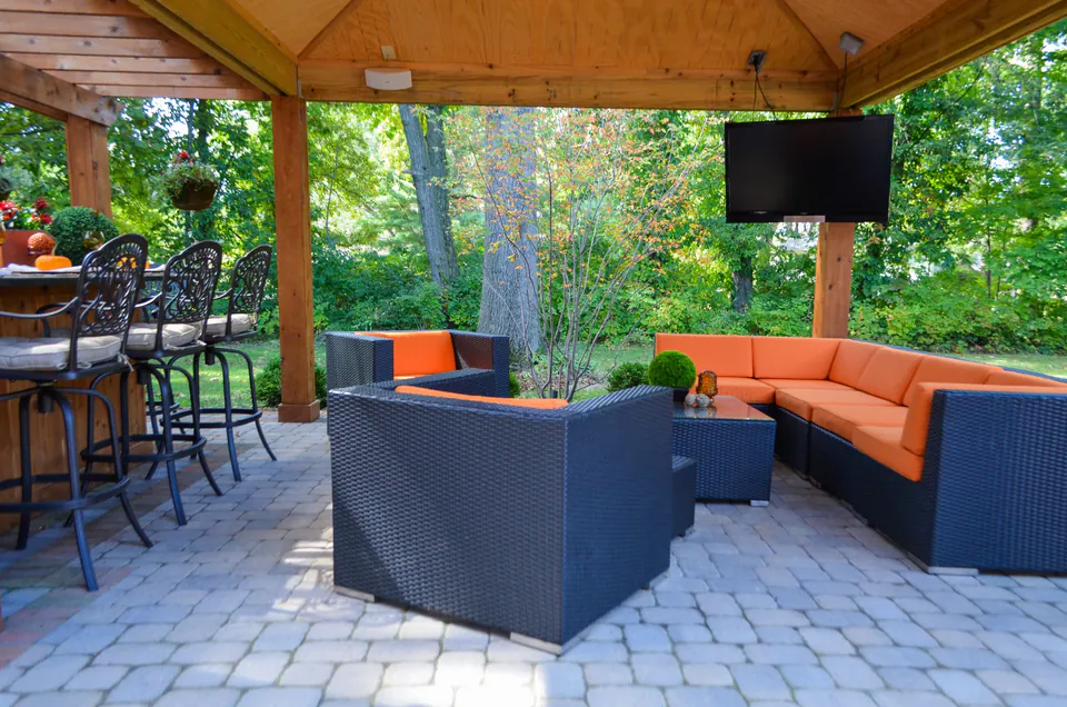 Covered outdoor room with grill bar, television, orange lounge seating, wood posts, and paver patio.